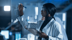 In the Research Laboratory Smart African American Female Scientist Wearing White Coat and Protective Glasses Writes Formula on Glass Whiteboard, References Her Tablet Computer